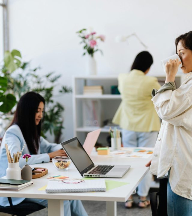 Three women in a modern office, working and interacting with laptops and plants around.